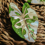 A close-up of Alocasia Earrings, shaped like a green and white leaf with metallic veins, resting on a woven, textured surface—perfect for those who love hand-crafted jewelry.