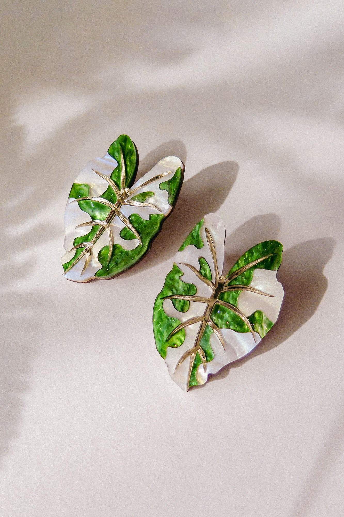 Two Alocasia Earrings shaped like monstera leaves with green and white enamel detailing are placed on a light surface, casting soft shadows—perfect for those who love lightweight jewelry.