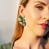 A woman with blonde hair wears a green ribbed top and a large Alocasia Earrings in a green and beige pattern. She is facing the camera in soft, natural light, showcasing the beauty of this hand-crafted jewelry.