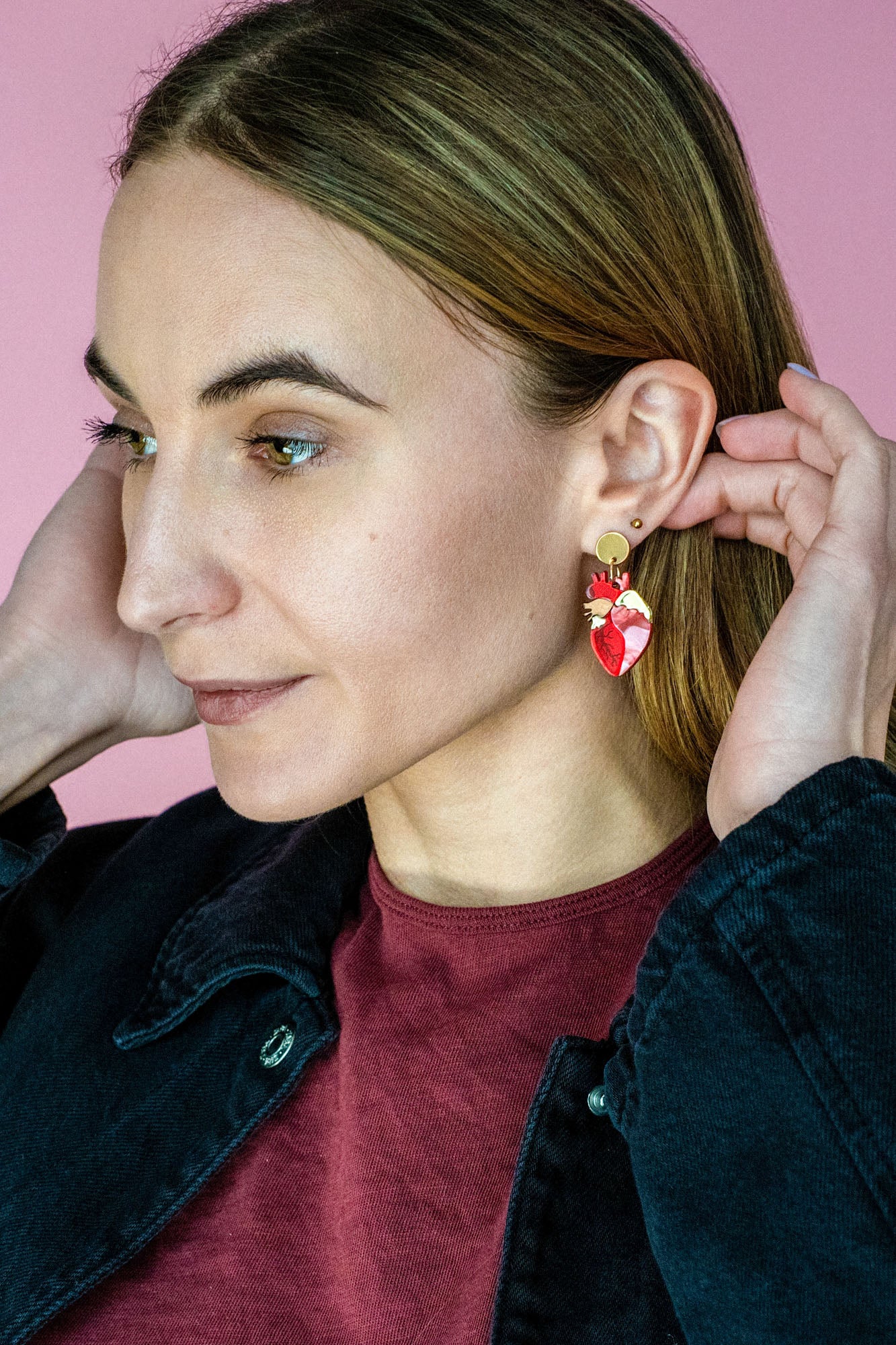 A woman with light brown hair wears a maroon shirt and black jacket, adjusting her unique acrylic earrings. She stands in front of a pink background, looking to the side with a slight smile.