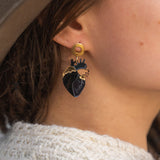 Close-up of a person wearing a gold and black Anatomical Heart Earring with gothic flair. The individual has light skin, brown hair, and is dressed in a beige hat and cream knitted sweater.