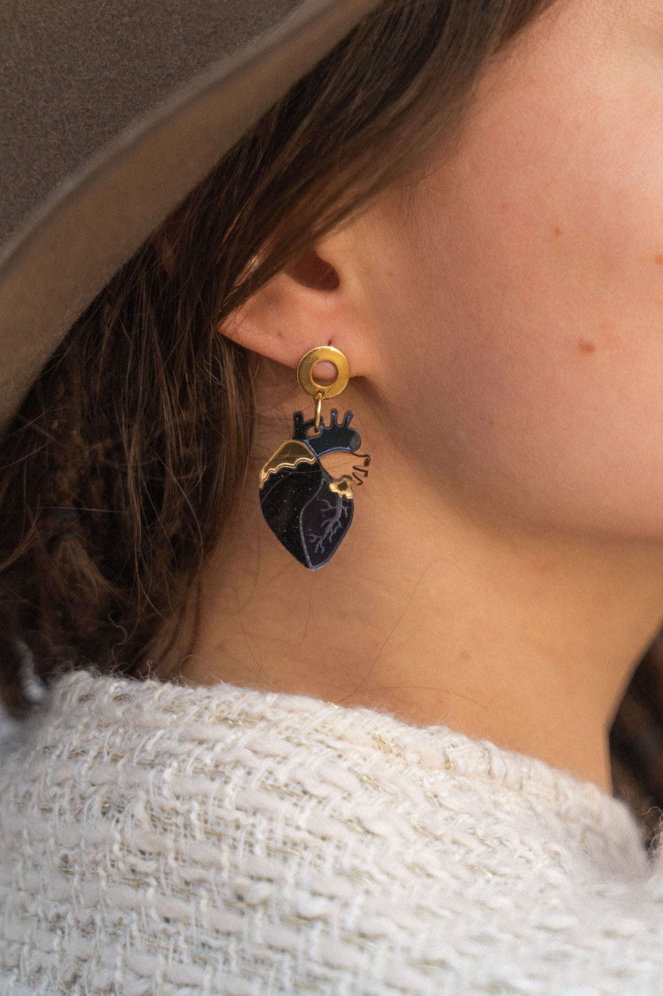 Close-up of a person wearing a gold and black Anatomical Heart Earring with gothic flair. The individual has light skin, brown hair, and is dressed in a beige hat and cream knitted sweater.