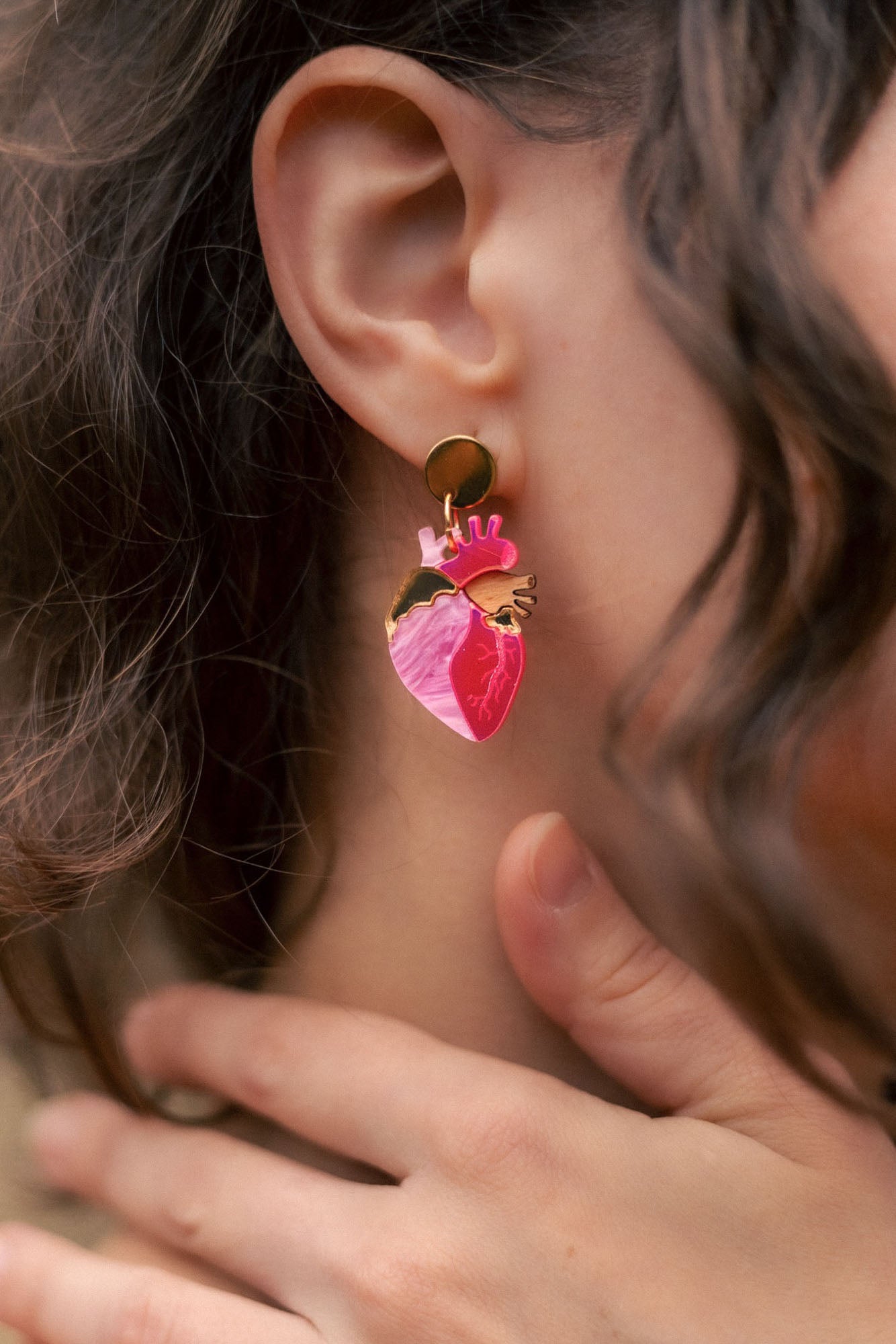 A close-up of a person’s ear wearing bold Anatomical Heart Earrings crafted from pink and gold holographic acrylic. Their curled brown hair partially frames the ear, while their hand gently touches their neck.