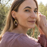 A woman with long light brown hair sits outdoors, smiling slightly as she looks over her shoulder. She wears a mauve blouse and handcrafted Bear Earrings made from premium acrylic glass, with sunlight and greenery in the background.