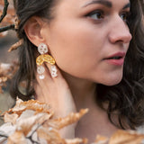 A woman with wavy brown hair wearing a cream top and boho chic dangle earrings stands among branches with dry, light brown leaves, gazing thoughtfully to the right.