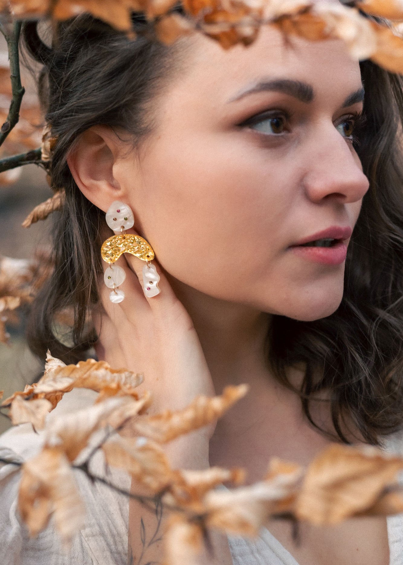 A woman with wavy brown hair wearing a cream top and boho chic dangle earrings stands among branches with dry, light brown leaves, gazing thoughtfully to the right.