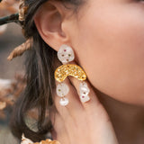 A close-up of a womans ear wearing unique boho chic dangle earrings with gold, pearl-like abstract shapes, and subtle rhinestones. Her hand touches her neck, surrounded by brown autumn leaves and branches.