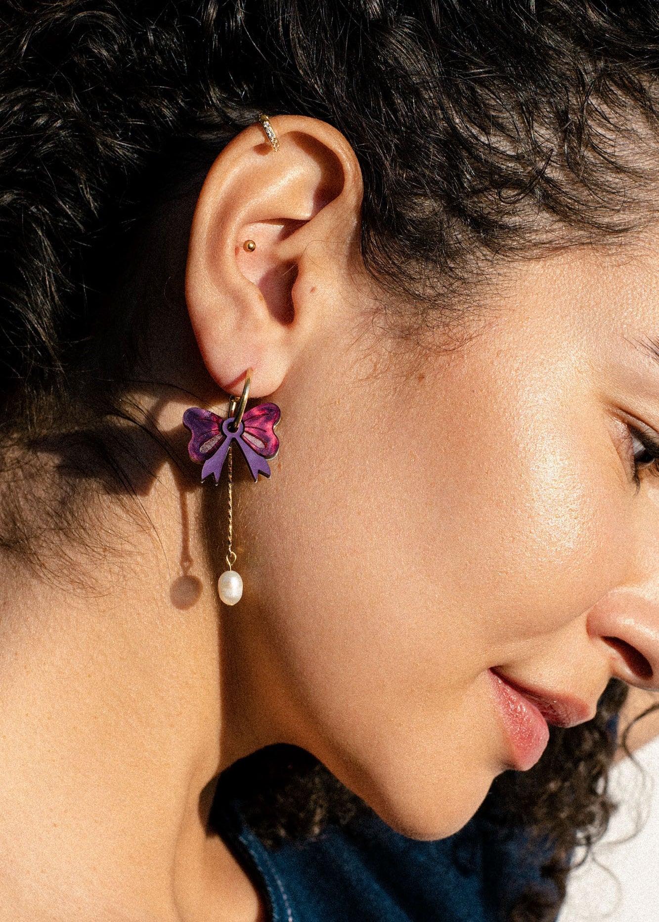 Close-up of a woman's ear with curly hair, wearing a gold ear cuff and an interchangeable dangle earring shaped like a purple bow with a dangling pearl. The side profile highlights her smooth skin and natural makeup.