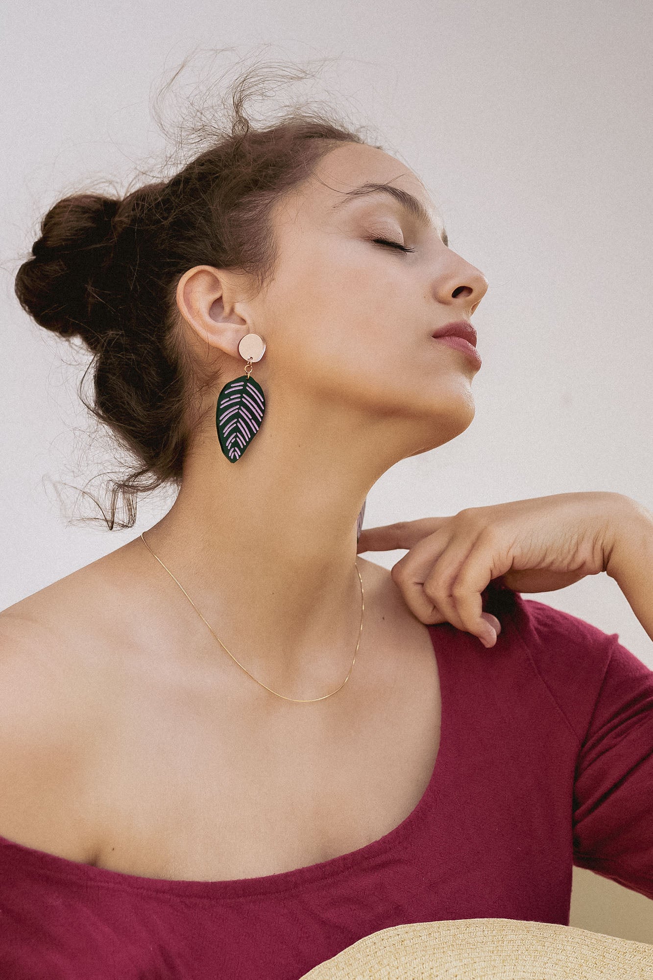 A woman with closed eyes and an elegant pose wears a burgundy off-the-shoulder top, a delicate gold necklace, and Calathea Ornata Earrings—handcrafted earrings inspired by tropical nature jewelry. Her dark hair is tied up as she holds a straw hat on her lap.
