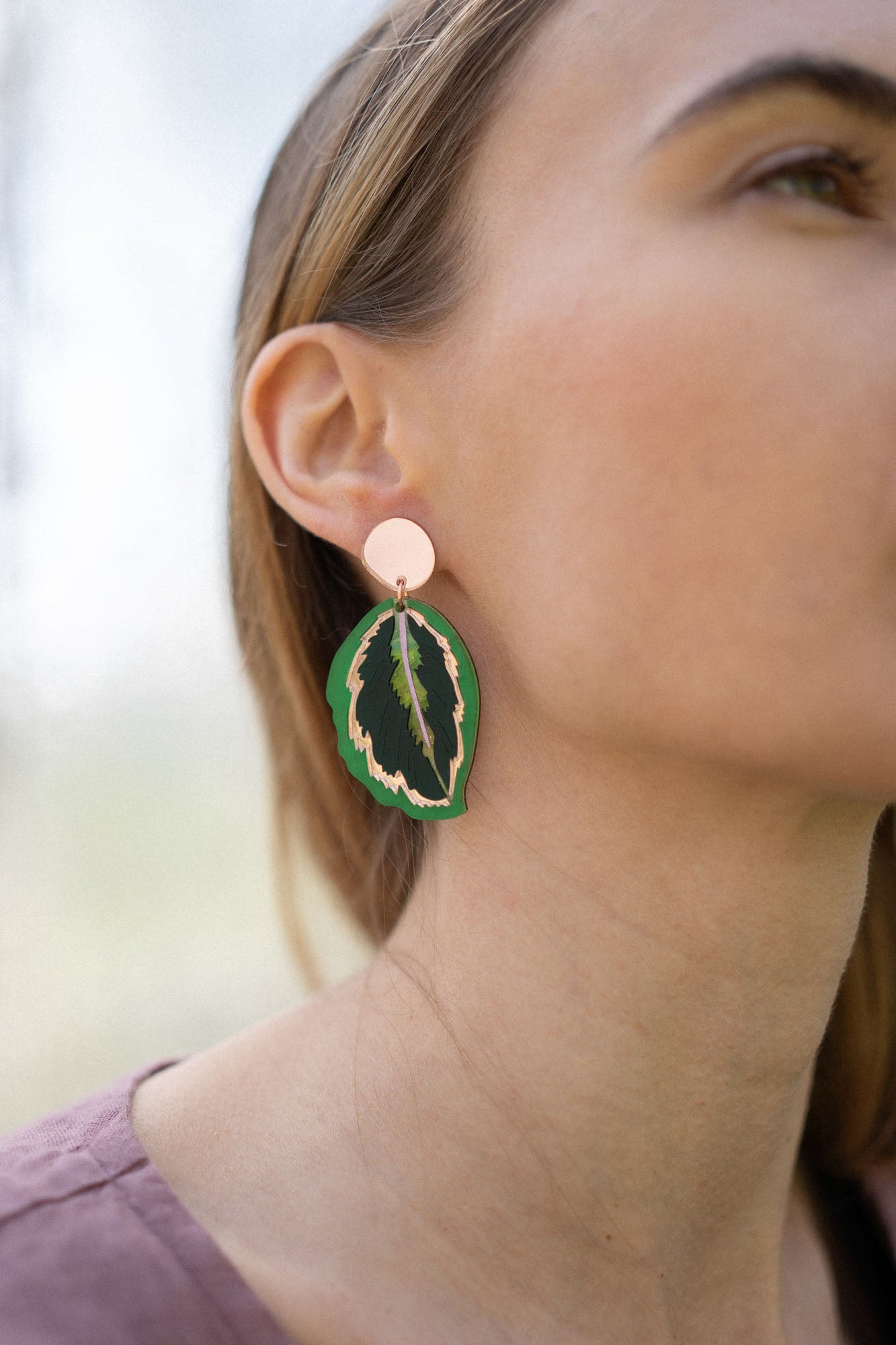 Close-up of a woman wearing Calathea Medallion Earrings—handcrafted statement jewelry in green and black with a gold edge. She has light brown hair, a mauve top, and only the right side of her face is visible.