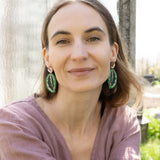 A woman with light brown hair, wearing a mauve blouse and Calathea Medallion Earrings, sits outdoors in sunlight, smiling softly at the camera. Blurred greenery and a glass panel create a serene backdrop for her nature-inspired jewelry.
