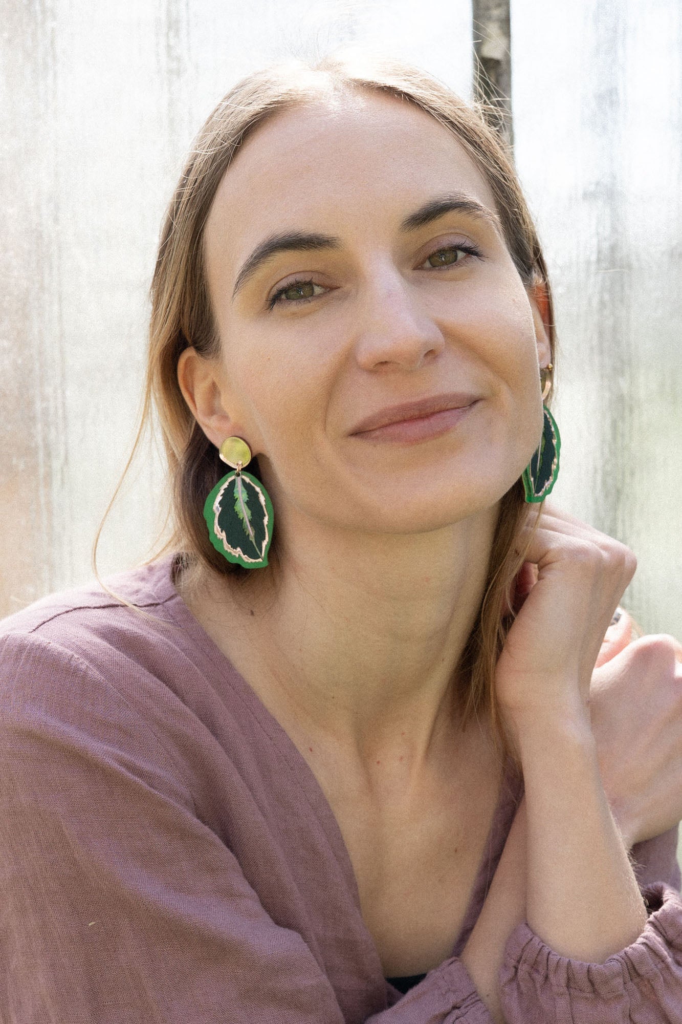 A woman with light brown hair, wearing a mauve blouse and green Calathea Medallion Earrings, sits by a sunlit window, smiling gently at the camera, her nature-inspired jewelry adding a unique touch to her look.