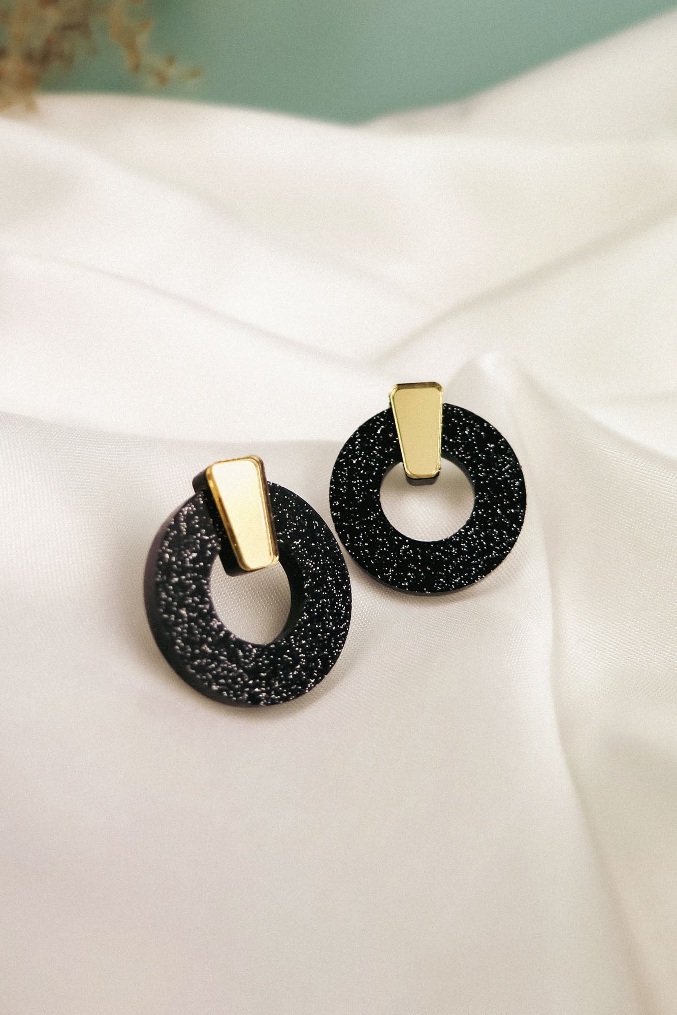 A pair of stylish Minimalist Circle Stud Earrings featuring black glittery acrylic glass hoops with gold rectangular accents, displayed on a white satin fabric background. Hypoallergenic for comfortable, all-day wear.