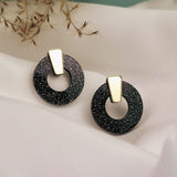 A pair of minimalist circle stud earrings in black acrylic glass with white speckles and gold triangular studs, displayed on white fabric with dried grasses in the background. Hypoallergenic for comfortable, everyday wear.
