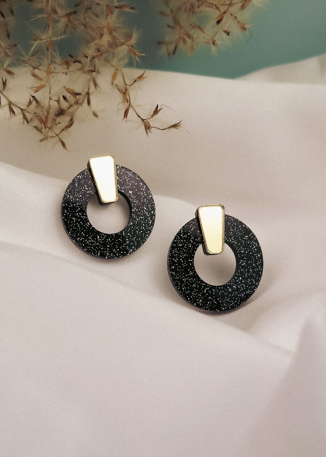 A pair of minimalist circle stud earrings in black acrylic glass with white speckles and gold triangular studs, displayed on white fabric with dried grasses in the background. Hypoallergenic for comfortable, everyday wear.