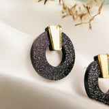 Close-up of minimalist circle stud earrings in black acrylic glass with silver glitter and a gold accent, displayed on a white fabric background with blurred dried plant details above. Hypoallergenic design ensures comfortable wear.