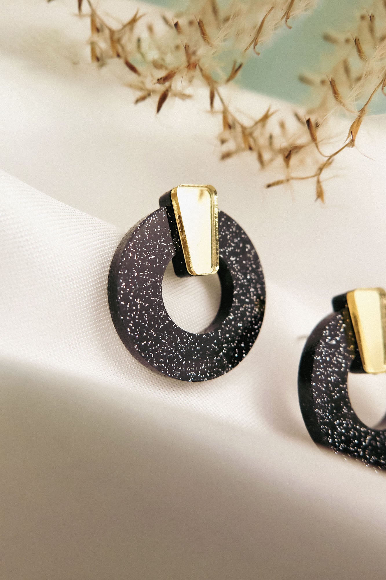 Close-up of minimalist circle stud earrings in black acrylic glass with silver glitter and a gold accent, displayed on a white fabric background with blurred dried plant details above. Hypoallergenic design ensures comfortable wear.