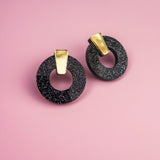 A pair of minimalist circle stud earrings, crafted from hypoallergenic acrylic glass with white speckles and gold rectangular accents, are displayed on a pink background.