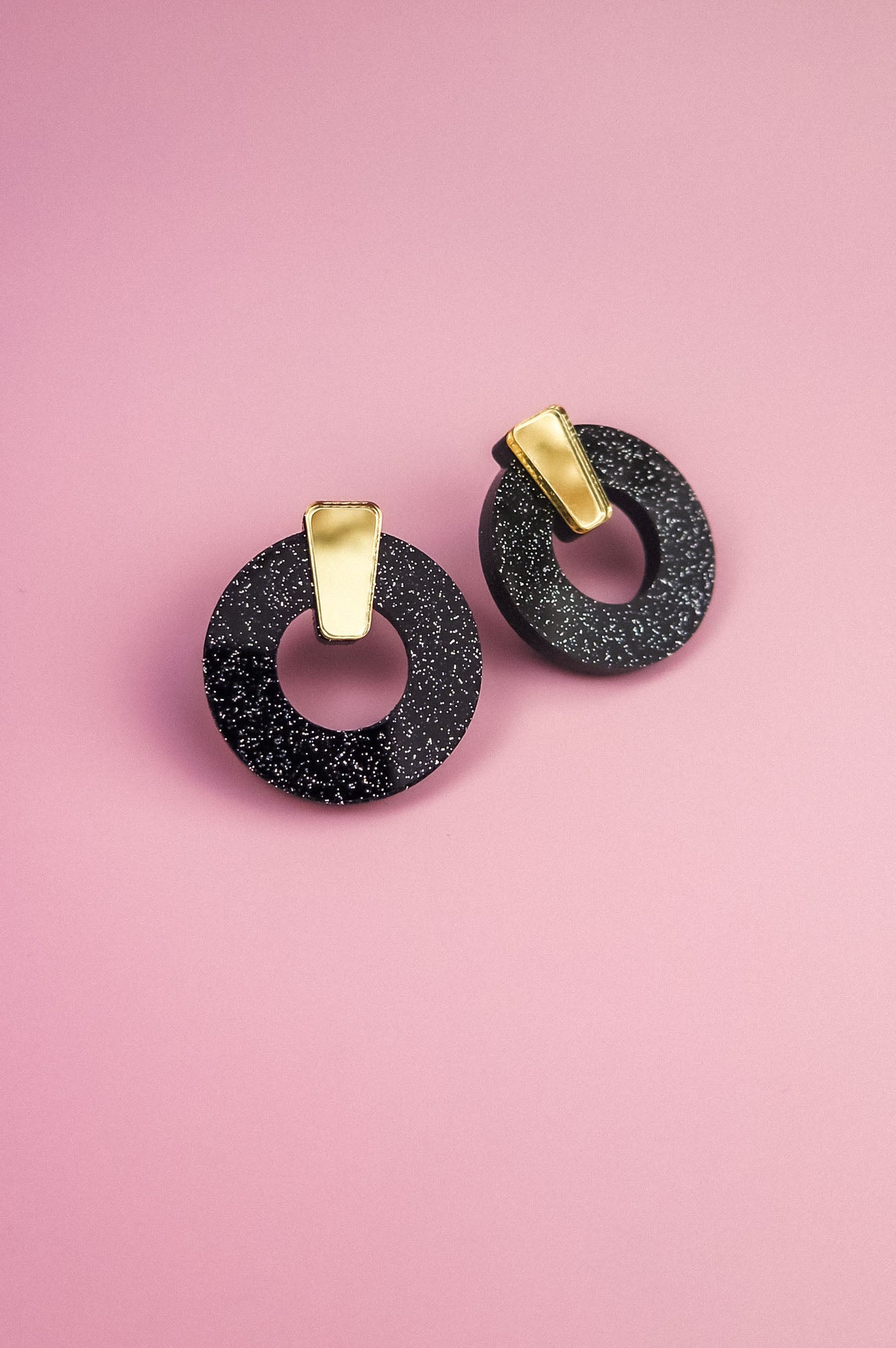 A pair of minimalist circle stud earrings, crafted from hypoallergenic acrylic glass with white speckles and gold rectangular accents, are displayed on a pink background.
