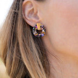 A close-up of a woman wearing Minimalist Circle Stud Earrings crafted from hypoallergenic materials, featuring a large, circular, multicolored gemstone with a gold accent, shown from the side against a blurred outdoor background.