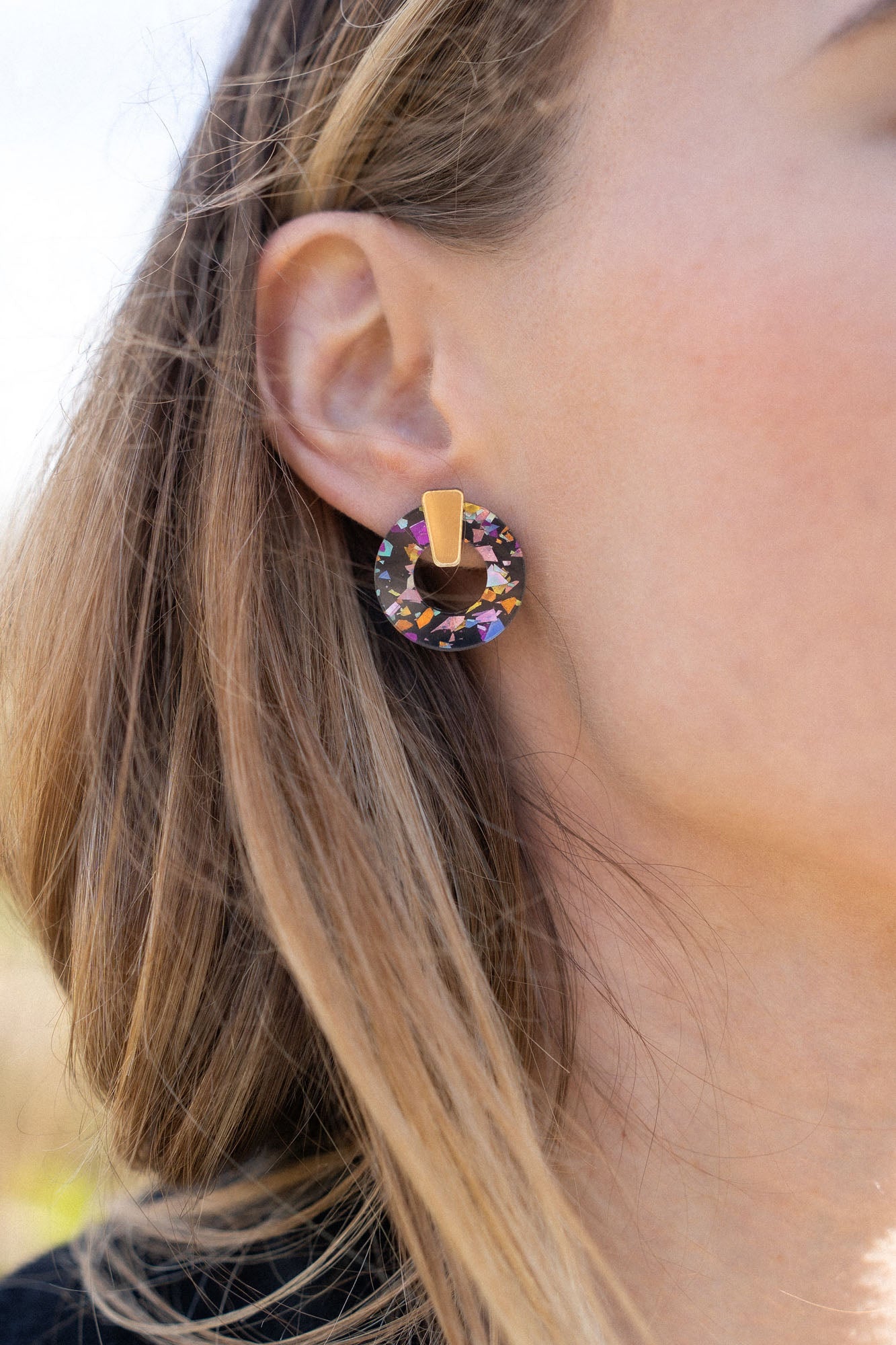 A close-up of a woman wearing Minimalist Circle Stud Earrings crafted from hypoallergenic materials, featuring a large, circular, multicolored gemstone with a gold accent, shown from the side against a blurred outdoor background.