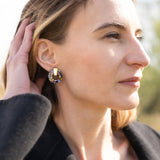 A woman with light brown hair touches her head while looking into the distance. She wears a black coat, a mauve top, and Minimalist Circle Stud Earrings that are hypoallergenic. The background is outdoors and softly blurred by sunlight.