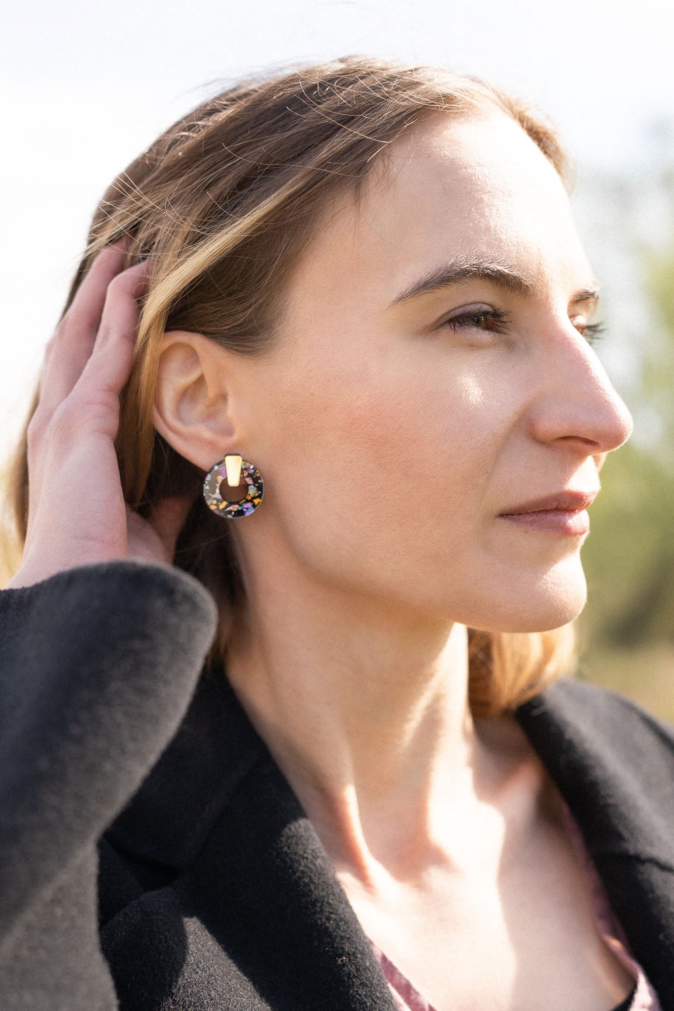 A woman with light brown hair touches her head while looking into the distance. She wears a black coat, a mauve top, and Minimalist Circle Stud Earrings that are hypoallergenic. The background is outdoors and softly blurred by sunlight.