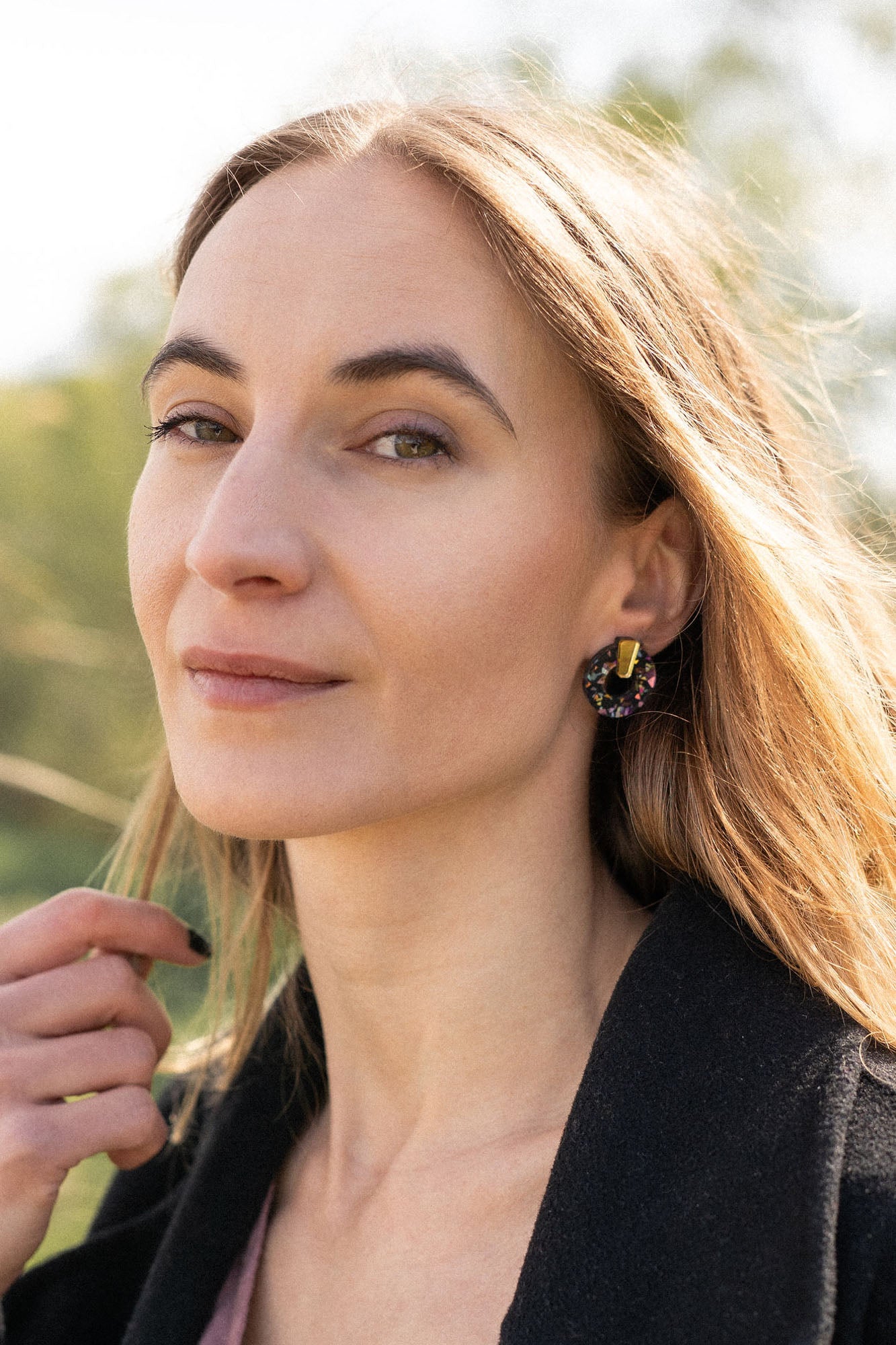A woman with long, light brown hair stands outdoors, wearing a dark coat, mauve top, and minimalist circle stud earrings. She lightly touches her hair and looks calmly at the camera against green foliage and a bright, sunlit sky.