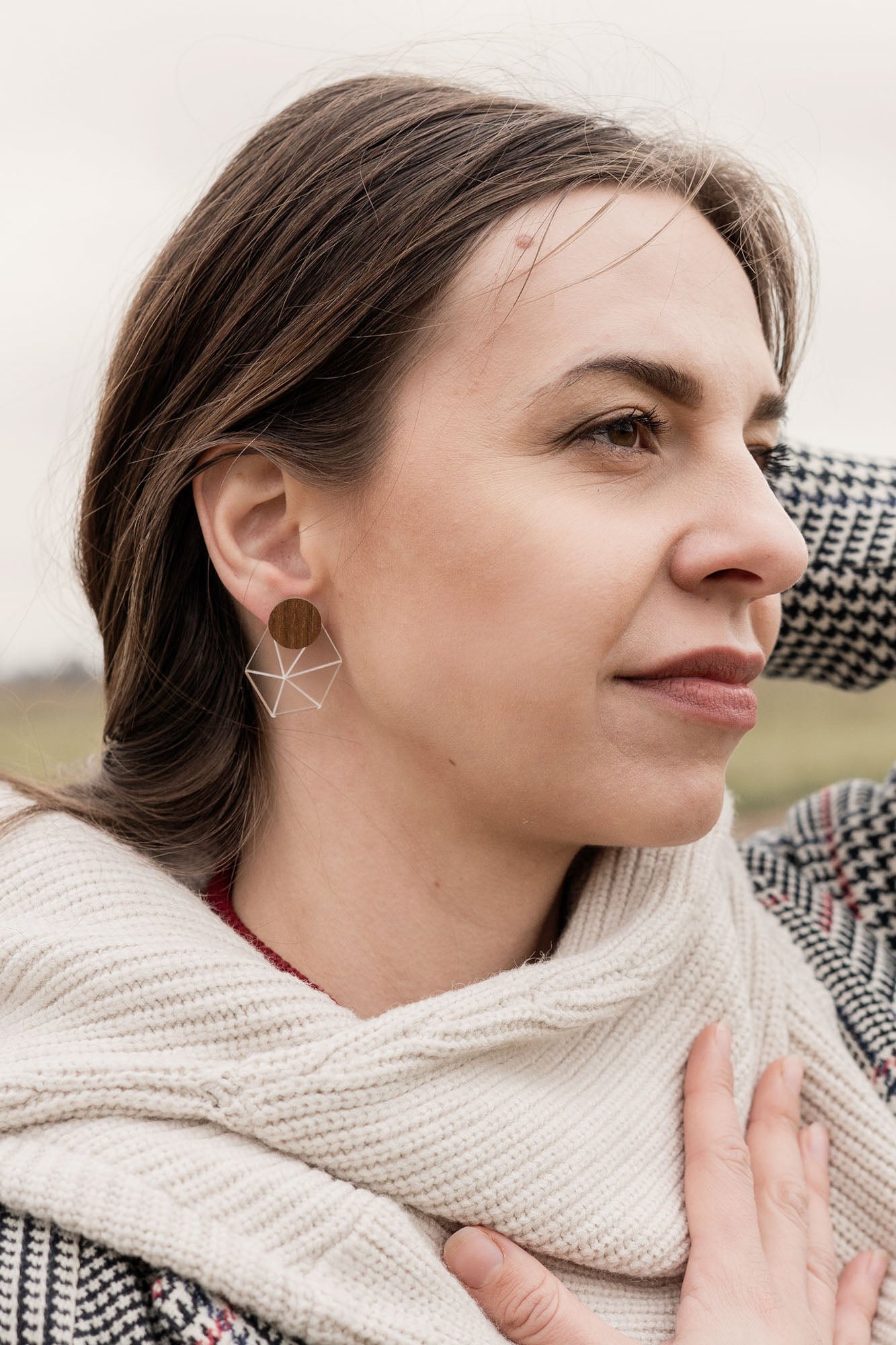 A woman in a houndstooth coat and cream scarf, embodying minimalist elegance, stands outdoors on a grassy, overcast day. She gazes thoughtfully into the distance, one hand on her chest and the other behind her head.