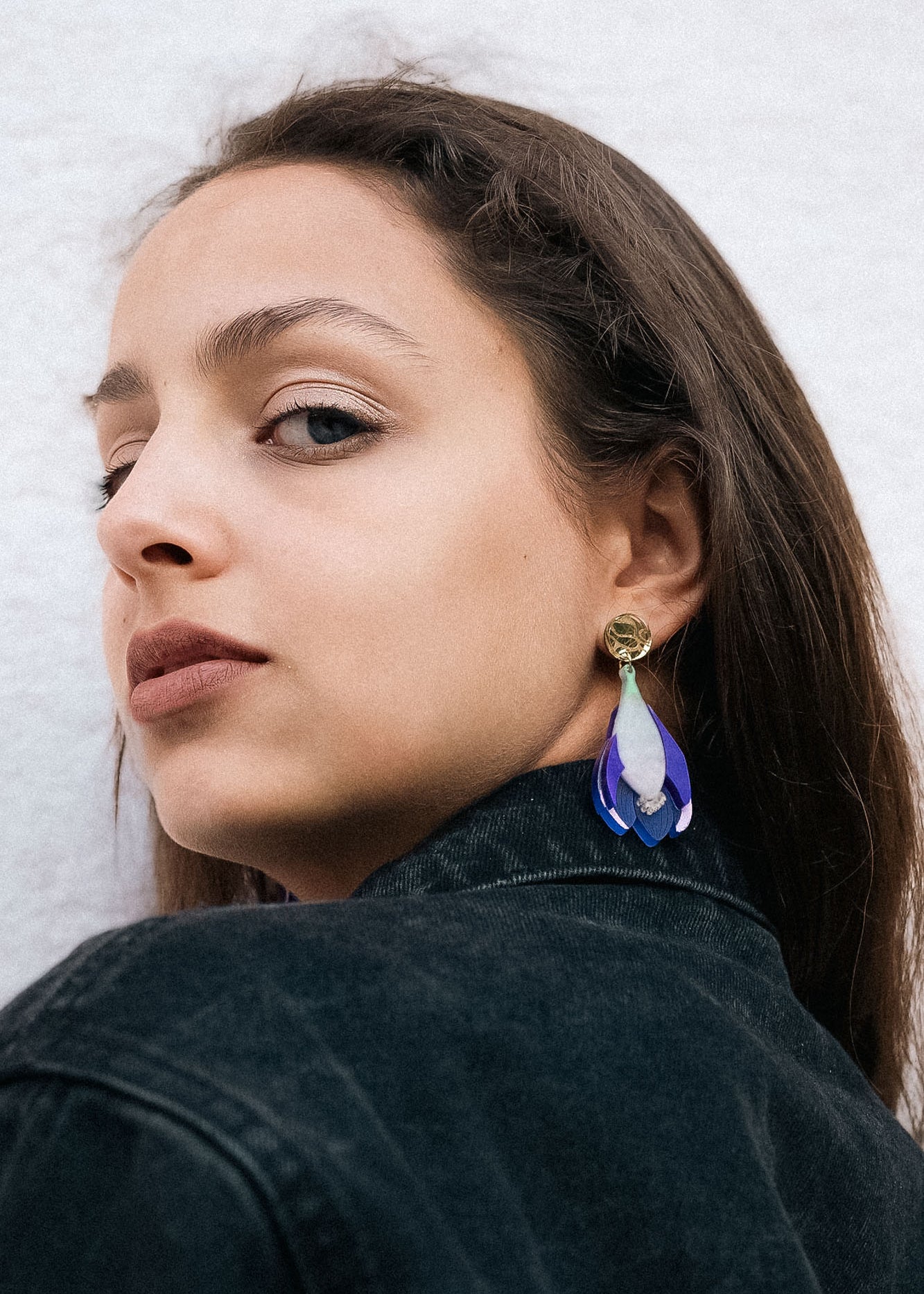 Woman with long brown hair wears a black denim jacket and handcrafted Crocus Flower Earrings, looking over her shoulder against a plain, light-colored background.