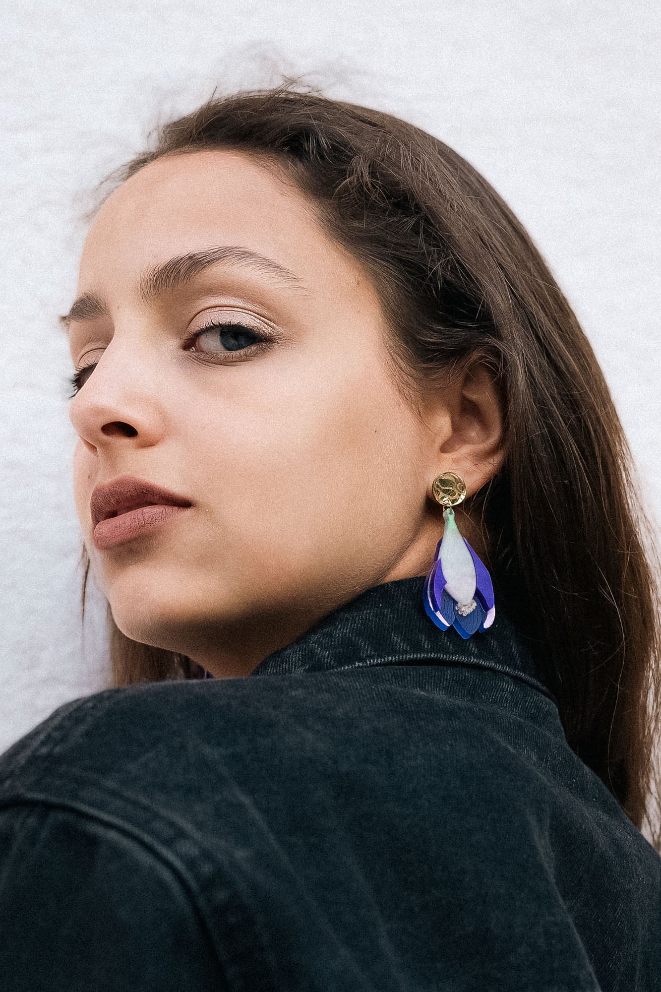 Woman with long brown hair wears a black denim jacket and handcrafted Crocus Flower Earrings, looking over her shoulder against a plain, light-colored background.