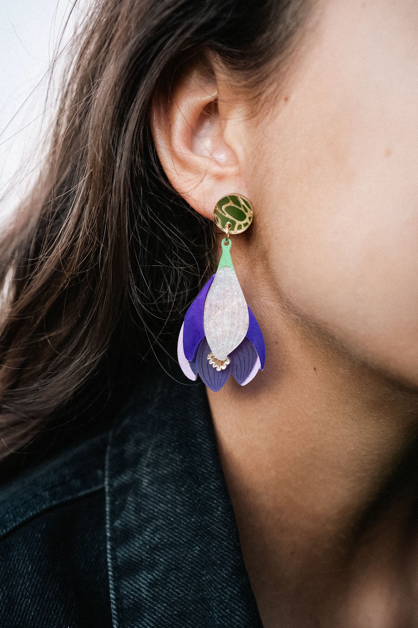 Close-up of a person wearing Crocus Flower Earrings—a handcrafted, statement accessory shaped like a flower petal in purple, white, and gold. The person has long brown hair and is dressed in a dark-colored top.