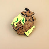 A playful brooch shaped like a dachshund lies on a plain beige background. With engraved details and bright yellow and green leaf-like accents, this wooden piece adds whimsy to any dog accessory collection.
