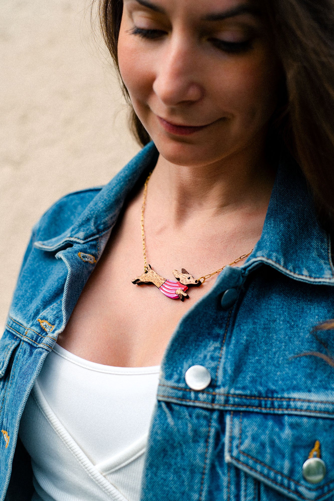 A woman wearing a denim jacket and white top looks down, showing a handcrafted gold necklace with a pendant shaped like a bird with outstretched wings and pink accents.