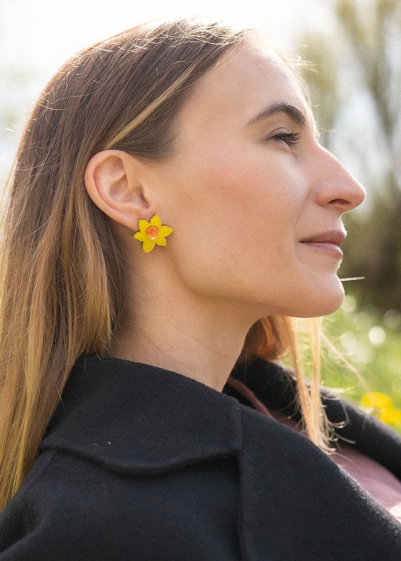 A stylish woman with long, straight hair reclines on grass outdoors, her unique accessories on display—Daffodil Stud Earrings adding charm as she looks peaceful, surrounded by blurred trees and greenery.