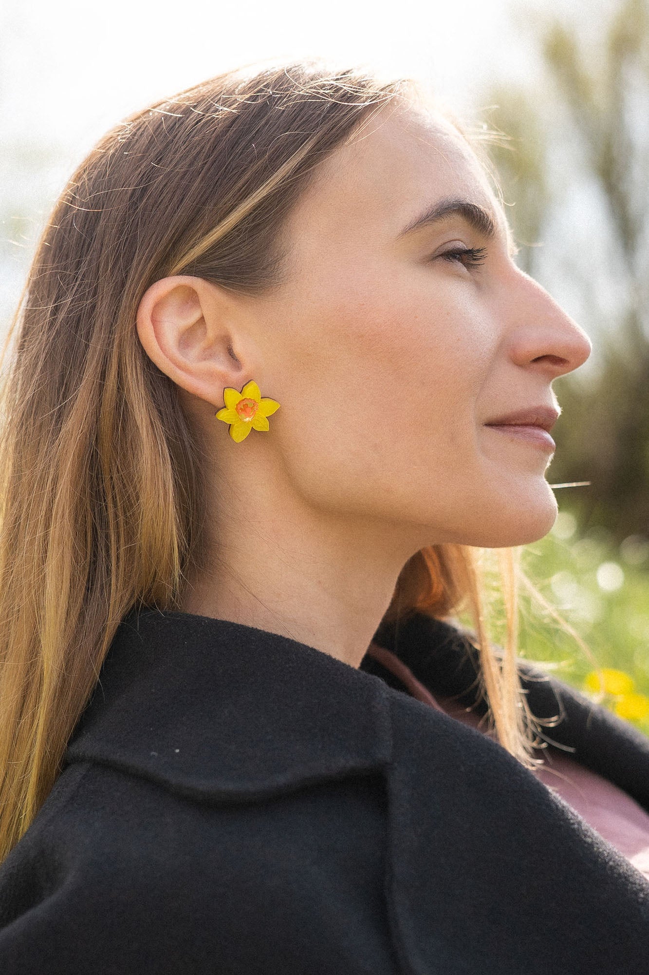 A stylish woman with long, straight hair reclines on grass outdoors, her unique accessories on display—Daffodil Stud Earrings adding charm as she looks peaceful, surrounded by blurred trees and greenery.