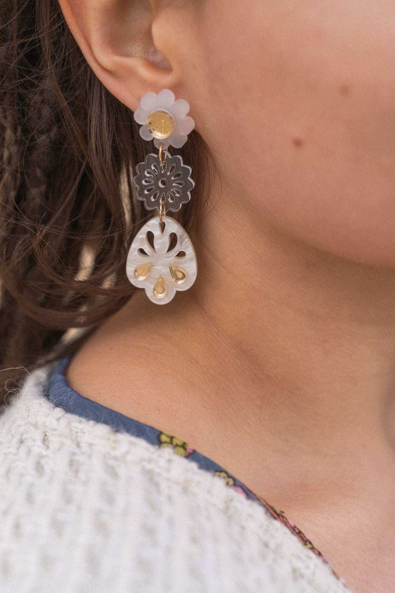 A close-up of a person wearing handcrafted, dangling earrings with flower and abstract shapes in translucent and gold tones, paired with a white textured sweater and a blue shirt with a floral pattern.