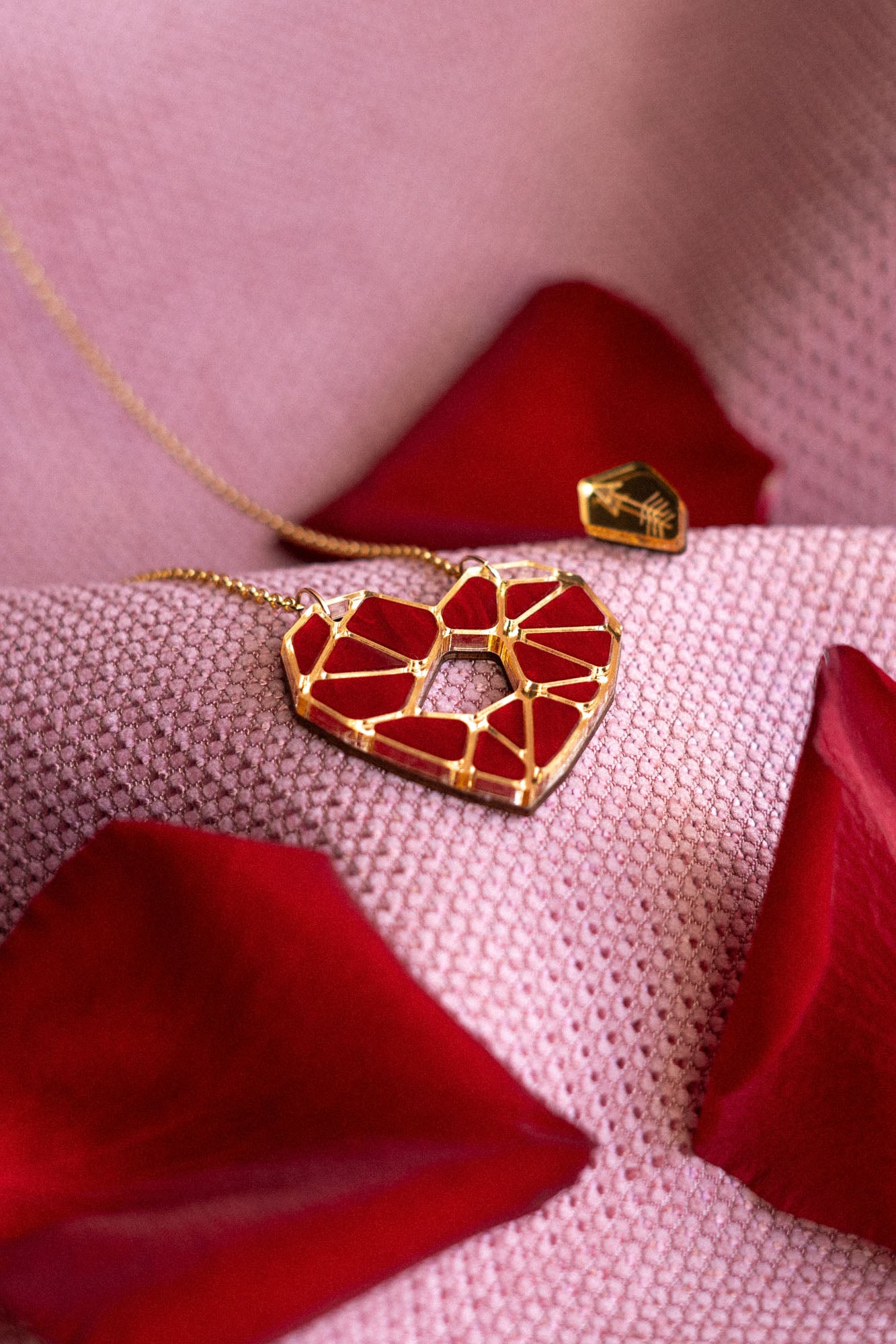 A handcrafted gold Fragment of Love Necklace with a geometric red heart pendant rests on soft pink fabric, surrounded by scattered red rose petals.