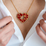 A close-up of a person in a white shirt, holding the collar open to reveal a handcrafted gold necklace with a geometric red heart pendant—an elegant Fragment of Love Necklace displayed around their neck.