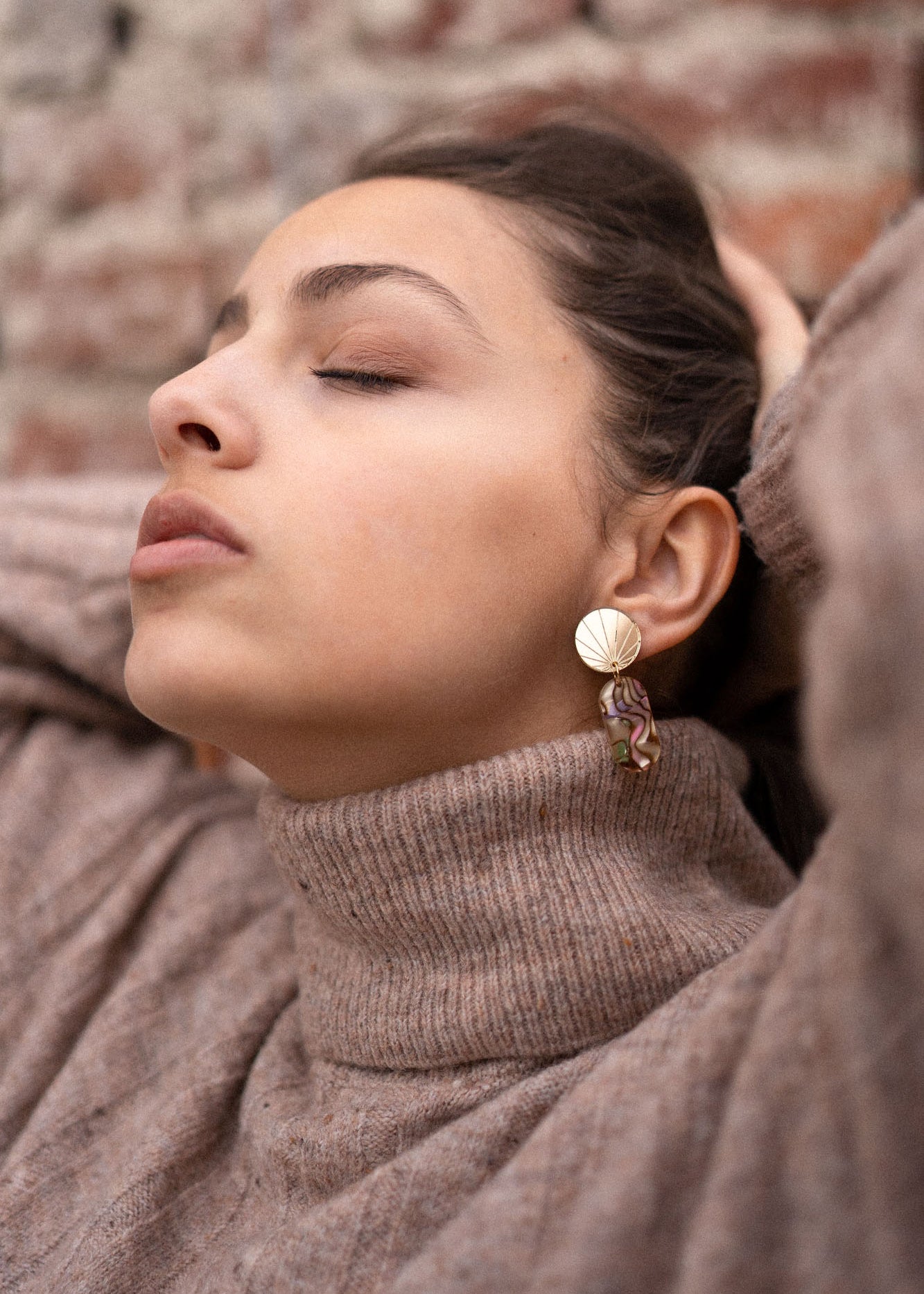 A woman wearing a beige turtleneck sweater and handcrafted gold earrings poses with her eyes closed, head tilted back, and hands resting behind her head in front of a brick wall.