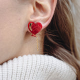 Close-up of a woman's ear wearing playful Heart Balloon Studs—red heart-shaped earrings with a small gold chain. Part of her face and blonde hair is visible, complemented by a white ribbed turtleneck sweater.