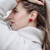 A woman in a cream-colored sweater stands outdoors, facing sideways with one hand holding back her long blonde hair. She wears playful Heart Balloon Studs, adding a romantic jewelry touch to her look. The background is blurry with trees and grass.