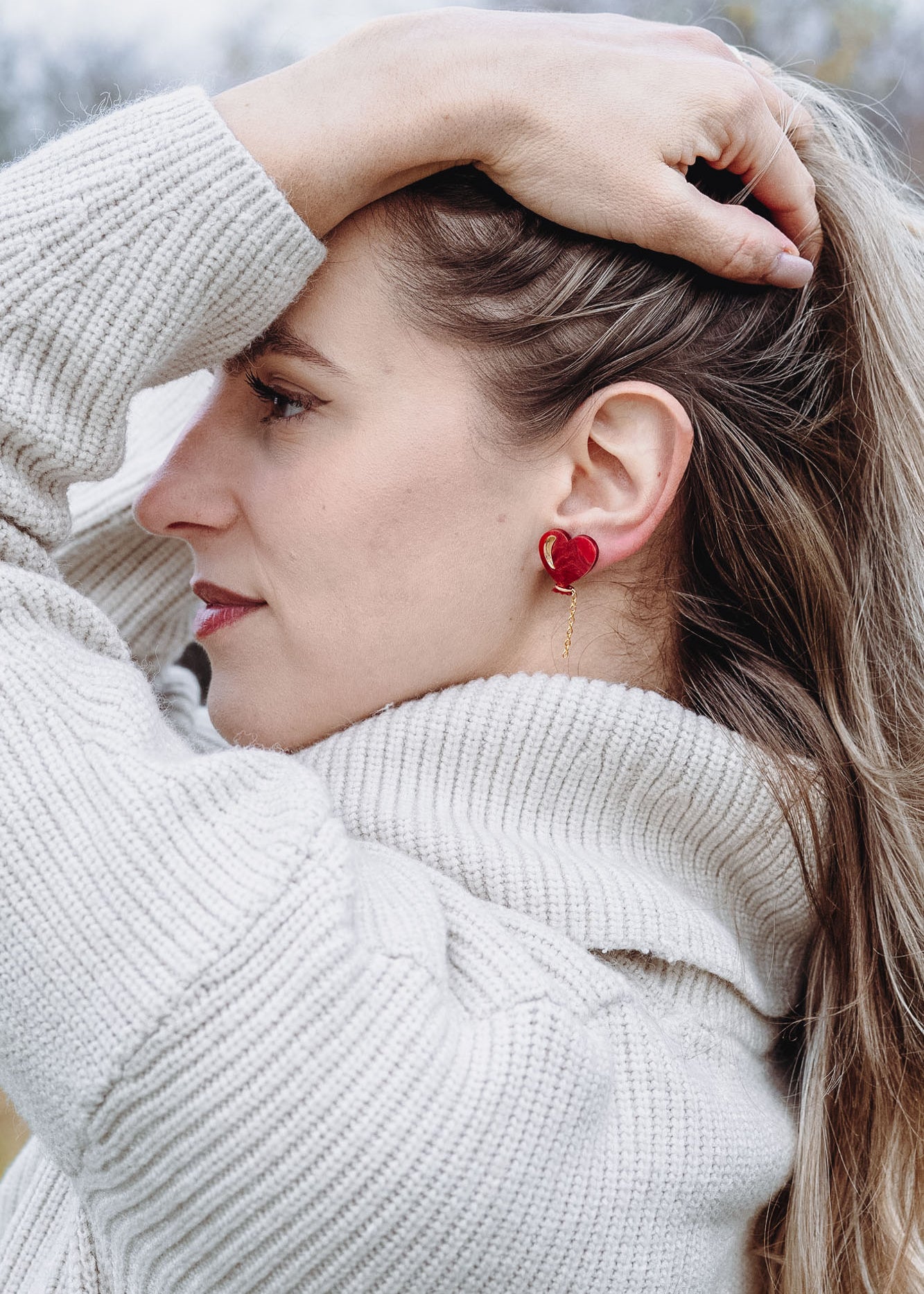 A woman in a cream-colored sweater stands outdoors, facing sideways with one hand holding back her long blonde hair. She wears playful Heart Balloon Studs, adding a romantic jewelry touch to her look. The background is blurry with trees and grass.