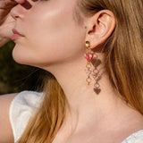 A woman with long brown dreadlocks and gold Heart Chain Earrings gazes to the left. She wears a cream-colored blouse and stands against a backdrop of pink curtains, highlighting soft pink tones throughout the scene.