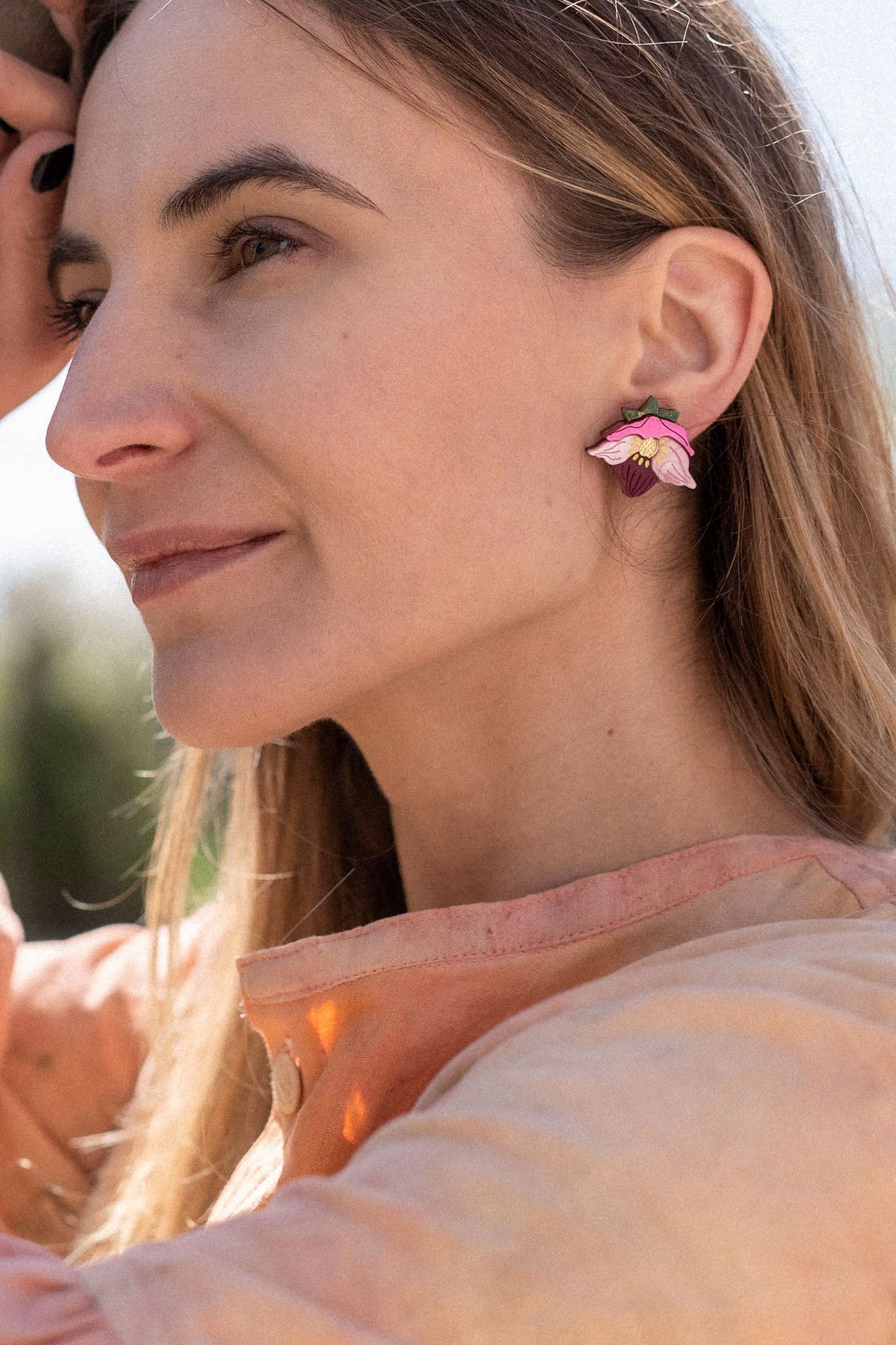A woman with long hair wearing a peach-colored blouse and handcrafted floral jewelry stands outdoors, gently touching a tree branch and smiling softly in natural sunlight.