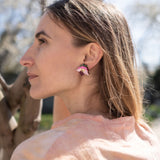 A woman with light brown hair stands outdoors near tree branches, wearing a light pink shirt and unique accessories—colorful, handcrafted floral jewelry that includes striking pink Hellebore Stud Earrings—as she gazes thoughtfully into the distance on a sunny day.