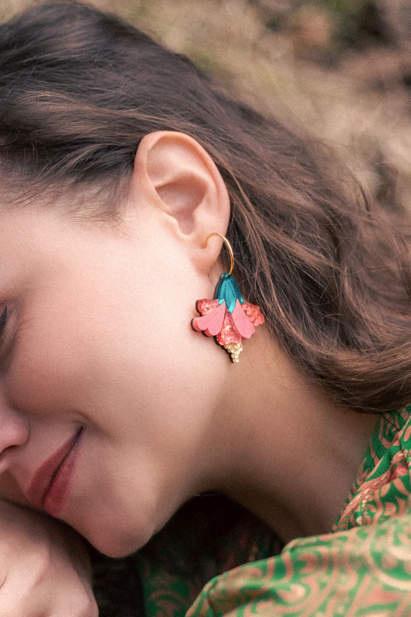 A woman with wavy brown hair wears vibrant Hibiscus Earrings—gold hoops adorned with pink, teal, and gold floral charms. She is smiling softly, her tropical statement green patterned clothing visible.