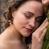 A woman with wavy brown hair and handcrafted Hibiscus Earrings gently leans her head and hand against a mossy tree, eyes closed, wearing an off-shoulder blouse—radiating a tropical statement in a serene forest setting.
