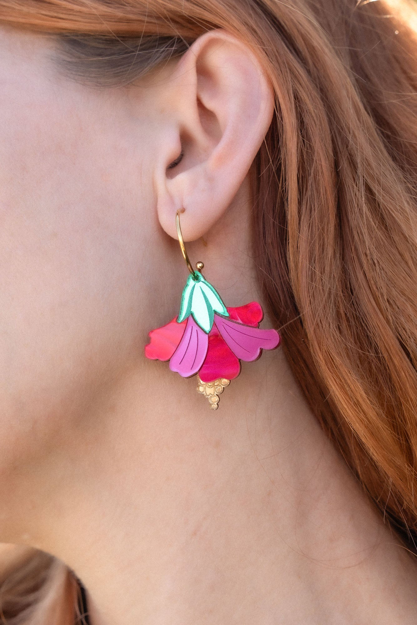 A close-up of a womans ear wearing handcrafted, petal-shaped Hibiscus Earrings in vibrant pink, teal, and gold tones. Her wavy, reddish-brown hair is partially visible, with tropical greenery softly blurred in the background.