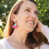 A woman with light brown hair smiles and looks upward, standing outdoors in sunlight. She wears a light long-sleeve top and handcrafted Hibiscus Earrings, with greenery and trees in the background.